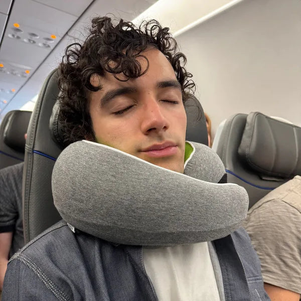 A traveler asleep in an airplane seat wearing the grey WANDER+ Pillow